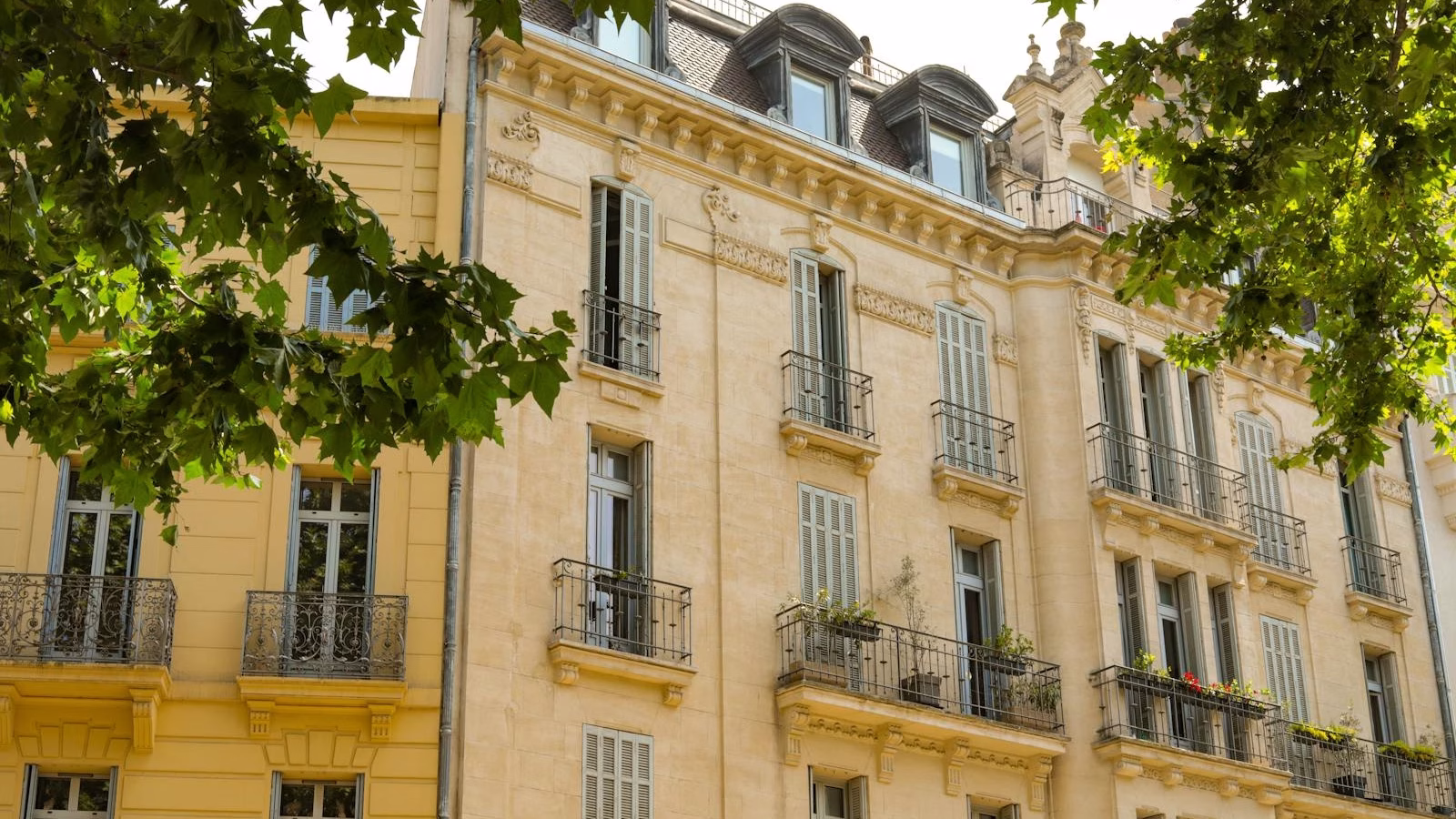 Elegant French-style architecture featuring balconies and detailed stonework under tree shade.